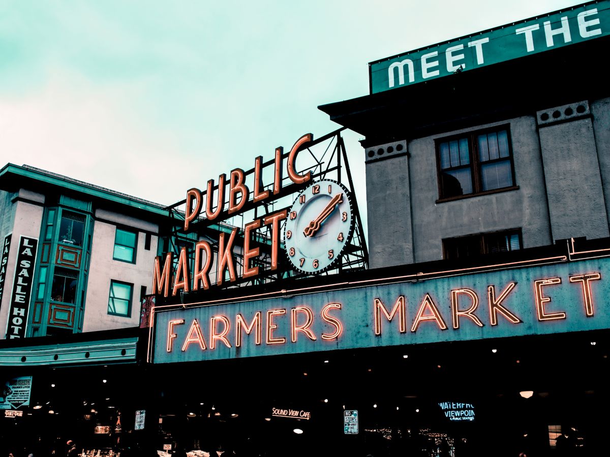 A public market sign with a clock and neon "Farmers Market" text, above a street scene with multiple buildings and overhead signage.
