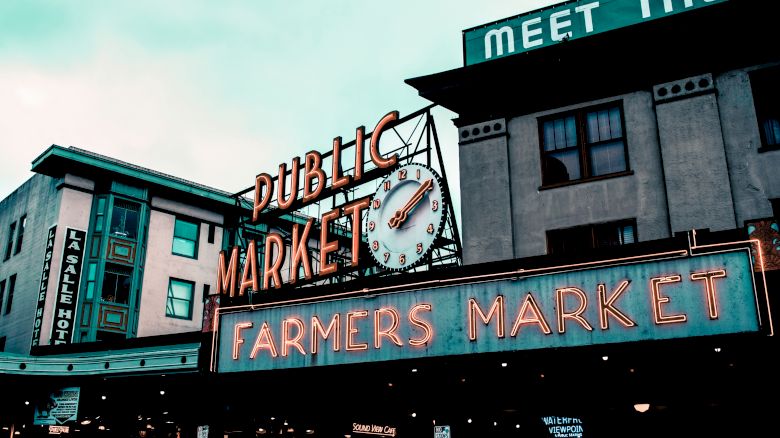 A public market sign with a clock and neon "Farmers Market" text, above a street scene with multiple buildings and overhead signage.