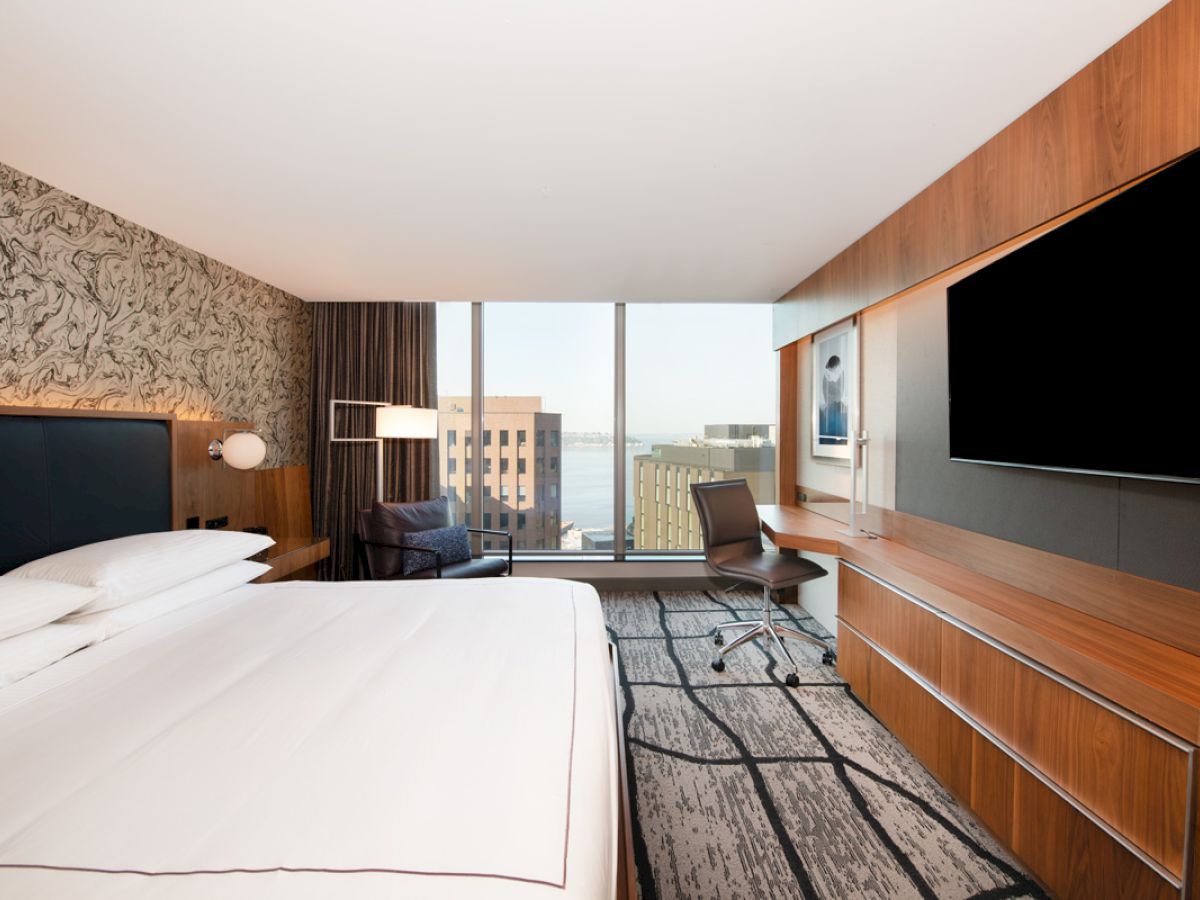 A modern hotel room with a large bed, wall-mounted TV, desk, chair, and a window view of city buildings and water.