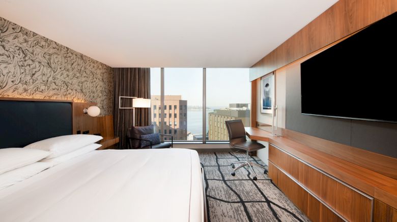 A modern hotel room with a large bed, wall-mounted TV, desk, chair, and a window view of city buildings and water.