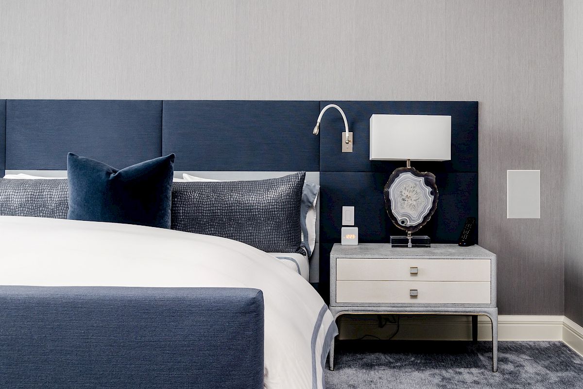 A modern bedroom with a blue upholstered bed, decorative pillows, and a nightstand with a lamp, on a soft carpeted floor.